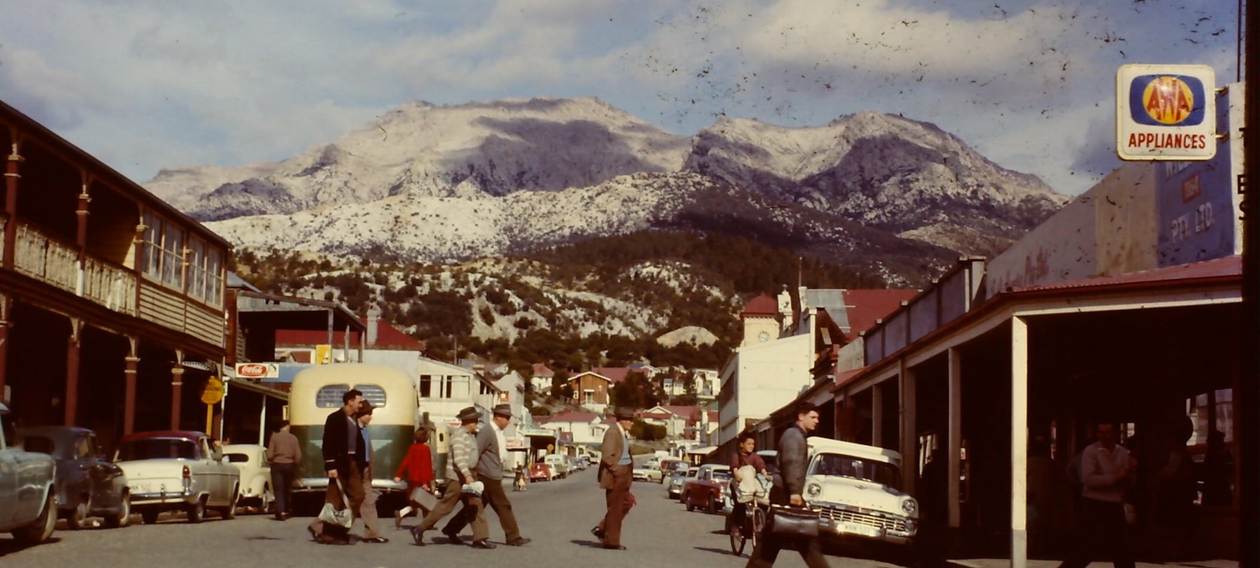 Shift Change. An old photograph of Queenstown's Orr Street. Vintage cars line the sides of the street and people cross the road wearing coats and hats, some carrying brief cases. In the background stands a snowy Mount Lyell. Credit Adrian Morgan.