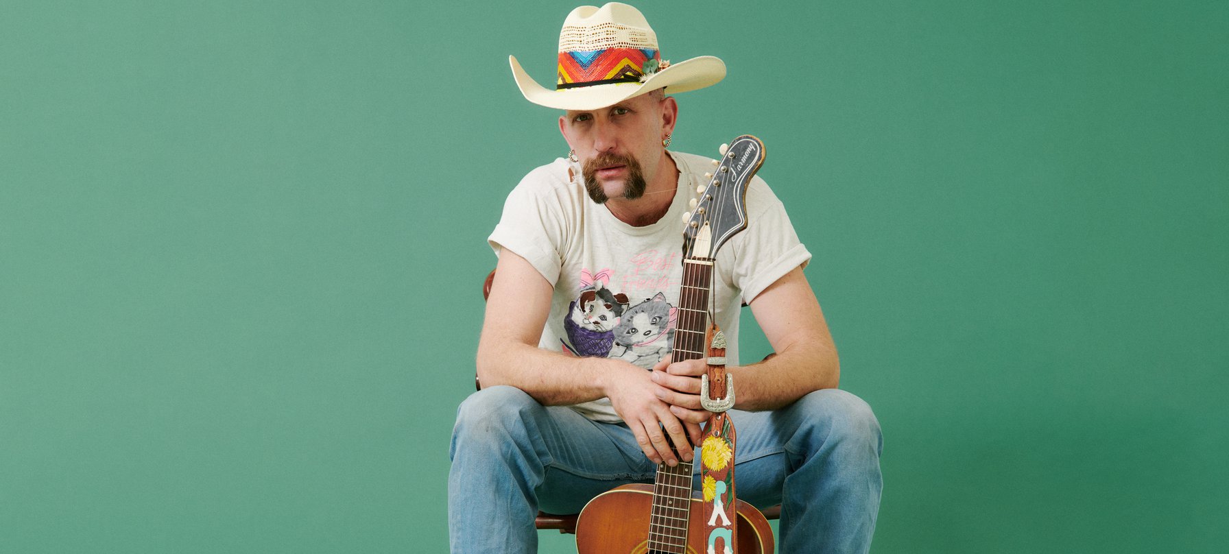 A photo of Andy Golledge sitting down, against a green background. Andy looks up from underneath a straw hat. He has a luscious moustache the shape of a downturned horse shoe, pale skin and thick gold hoop earrings. Credit Alex Wall﻿﻿.