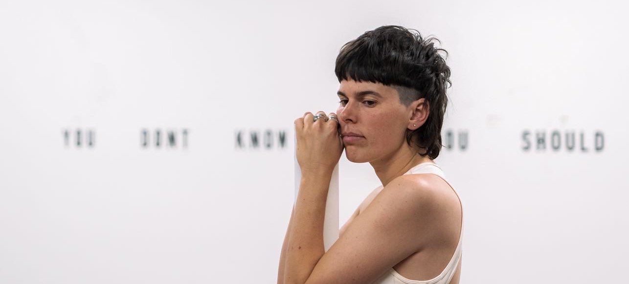 A photo of Ashleigh Musk side-on with a white background with blurred black words in a line along the wall. Ashleigh leans on a plinth, looking contemplatively into the distance. Ashleigh has pale skin and a luscious brown mullet. Credit Ivan Trigo Miras.