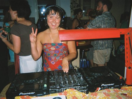 DJ Buoy Ambler stands behind a DJ deck at a house party in a kitchen, making a peace gesture with their hands and sticking their tongue out playfully.