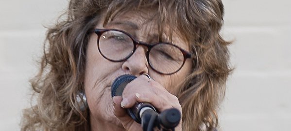 A photo of Aunty Cheryl Mundy wearing, Aunty Cheryl has her eyes closed as she sings into a microphone. Hey curly brown hair is flowing in the wind as she performs. Credit