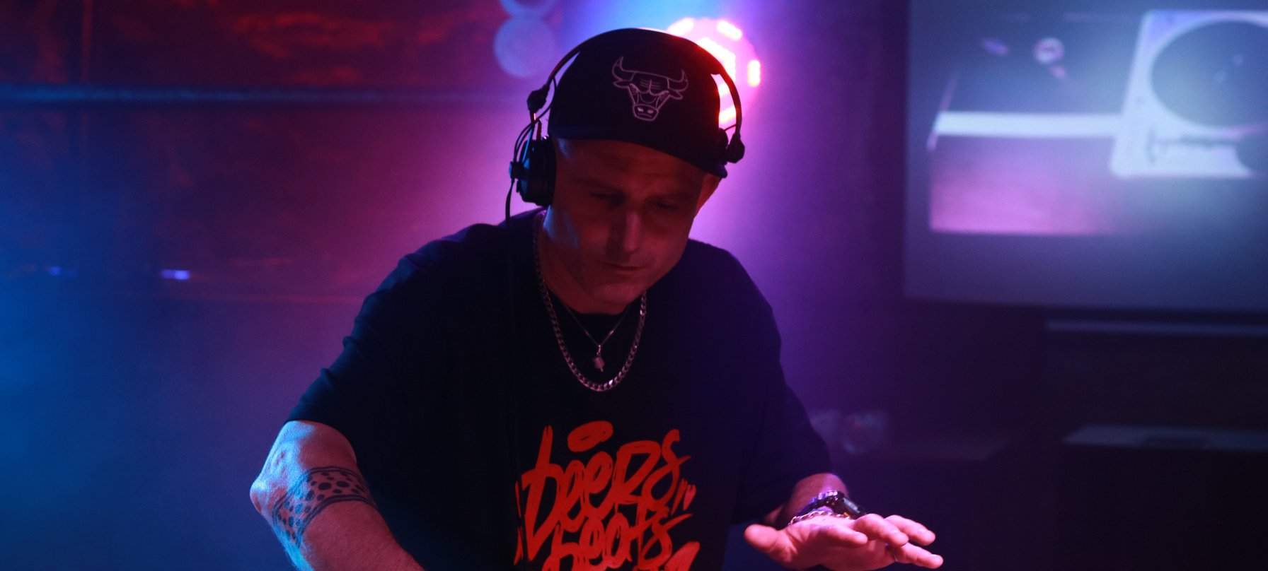 A photo of DJ Thensum leading over a DJ deck in a dark space with red and blue lights in the background. DJ Thensum has pale skin and is wearing a black cap cap and black and red tshirt. Credit Kishka Jensen.