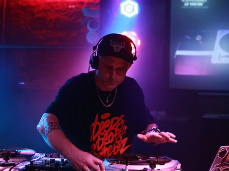 A photo of DJ Thensum leading over a DJ deck in a dark space with red and blue lights in the background. DJ Thensum has pale skin and is wearing a black cap cap and black and red tshirt. Credit Kishka Jensen.