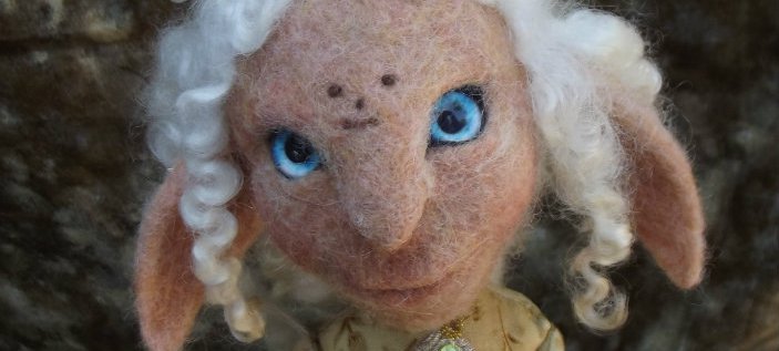 A photo of Billie Lee Martin's felt work. The work is a felt creature with human-like facial features, goat-like ears, tall twisting white horns and curly white hair. The figure is wearing and green and sequinned, satin garment. Credit Billie Lee Martin.