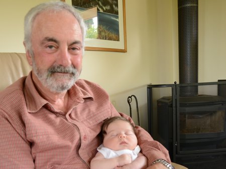 A photo of Lou Rae sitting in an armchair, cradling his newly-born granddaughter, Gracie. Lou, with pale skin and short white hair and beard smiles up at the camera while Gracie sleeps in his arms. Credit courtesy of artist.