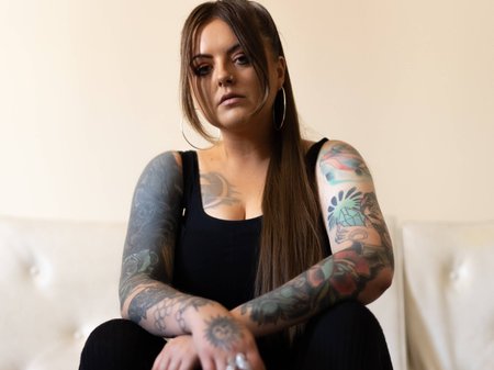 A photo of DENNI. DENNI is looking down at the camera, with dark brown hair falling either side of their face and large hoop earrings. DENNI has tanned skin and two colourful tattoo arm sleeves. Credit TBC.