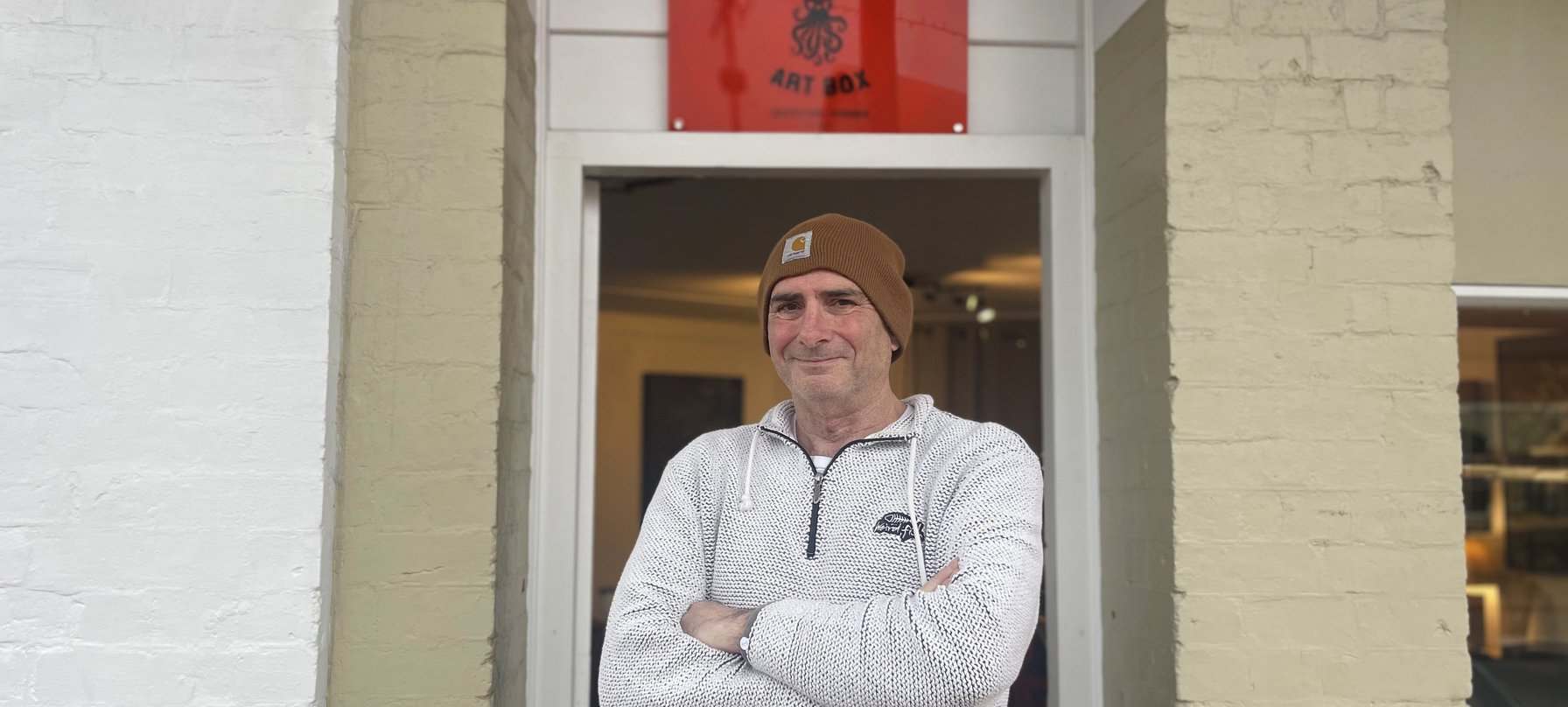 A photo of Doc Doran standing outside Empire Art Box in Queenstown with his arms crossed and a smile on his face.