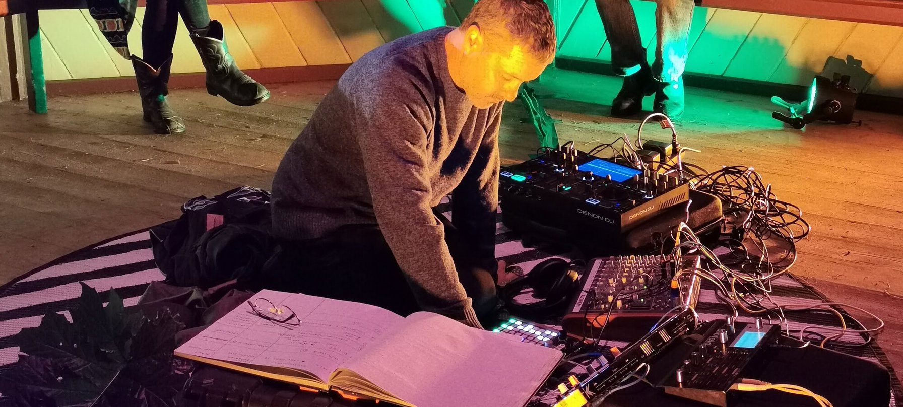 A photo of Dylan Martorell kneeling on the ground and surrounded by a circle of road cases, sound mixing boards and a lot of cords. Dylan is tinkering with a mixing board as audience members watch on. Credit Chuan Lim.