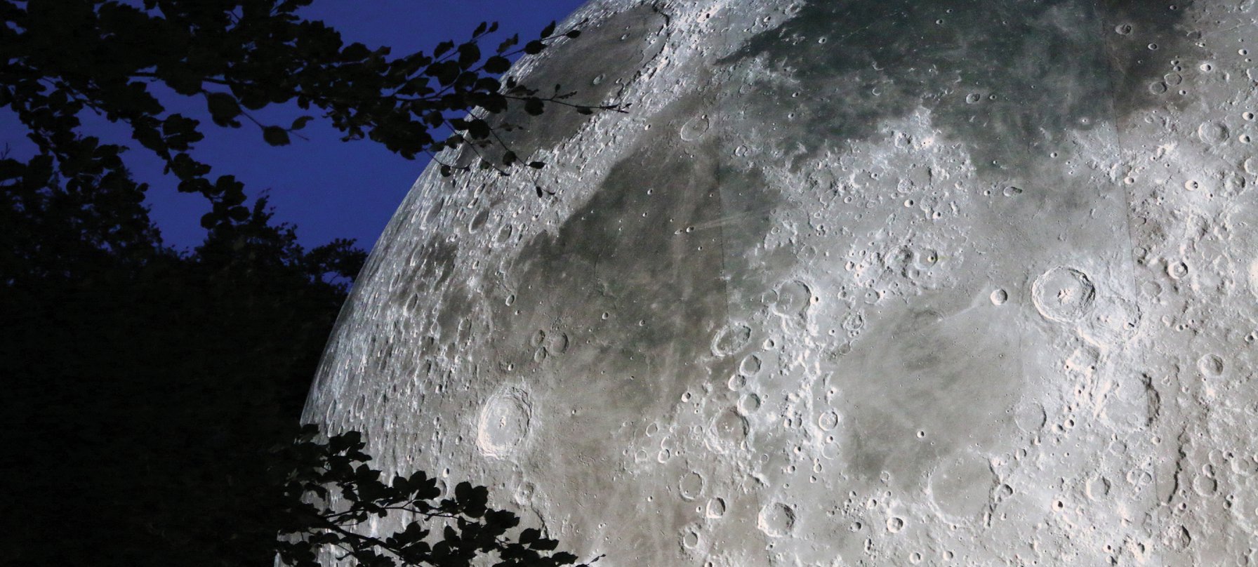 Earthshine image. A close-up photo of the Earthshine artwork. A large moon fills the space with its huge round body and shadowy face. A brilliant glow shines from the moon, showing its craters and hills. Credit Luke Jerram.