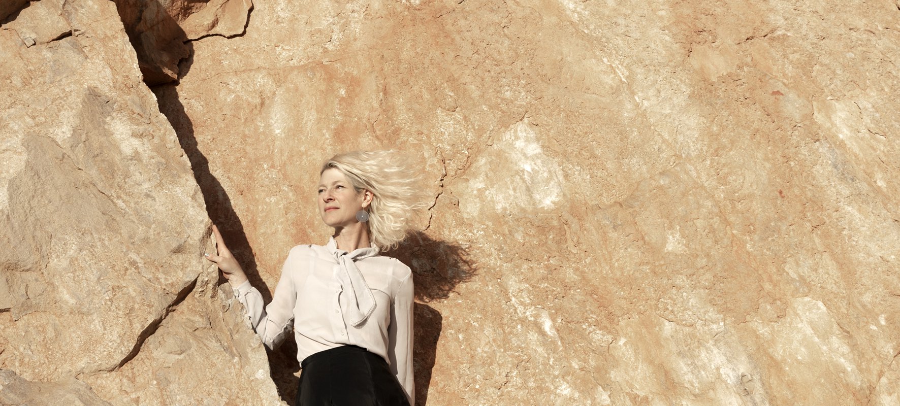 A photo of Elliat Rich standing in a crevice of a rock-face exposed as part of an extractive mining practice. Wind is blowing through Elliat's short blonde hair as they look into the distance. Credit Martina Capurso.