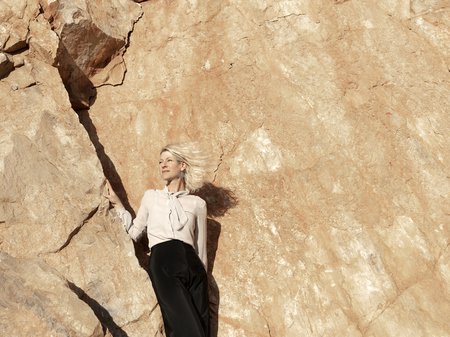 A photo of Elliat Rich standing in a crevice of a rock-face exposed as part of an extractive mining practice. Wind is blowing through Elliat's short blonde hair as they look into the distance. Credit Martina Capurso.
