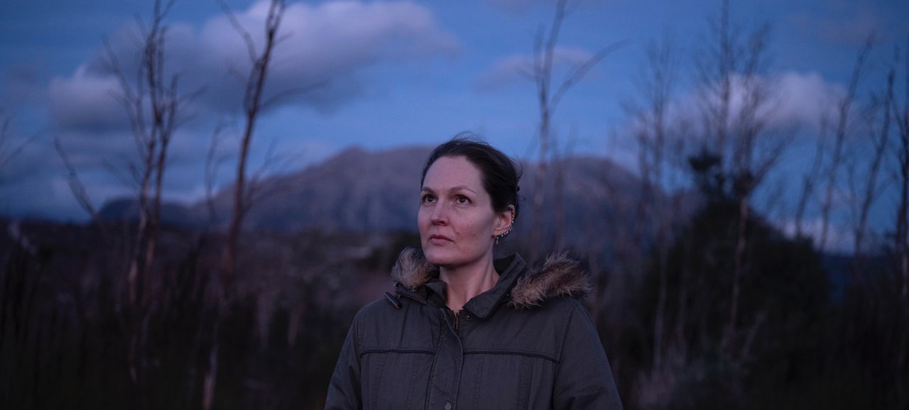 A photo of Emilie Llewellyn dressed in a warm jacket and standing amongst bare trees. In the distance is a cloudy blue sky above a mountain. Credit Sarah Rhodes.