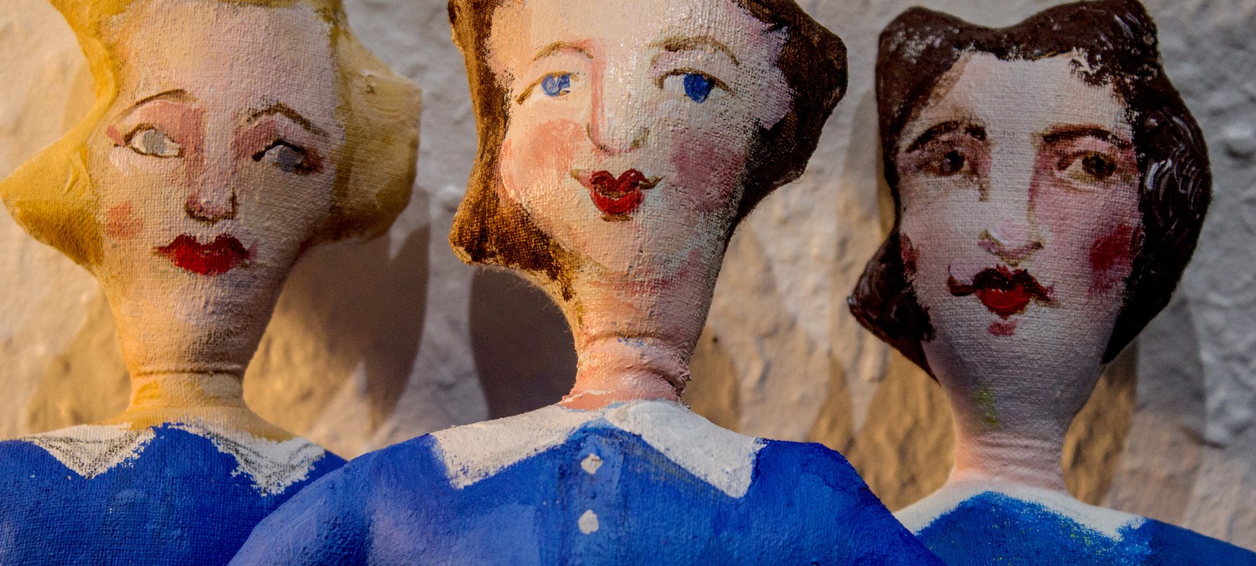 A photo of Grace Garton's work featuring three textile figures painted with blue dresses and 1940's-style feminine hair-dos. The figures all have painted faces with red cheeks and red lipstick. Credit Grace Garton.