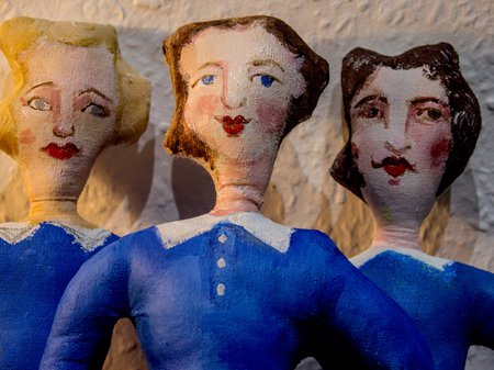 A photo of Grace Garton's work featuring three textile figures painted with blue dresses and 1940's-style feminine hair-dos. The figures all have painted faces with red cheeks and red lipstick. Credit Grace Garton.