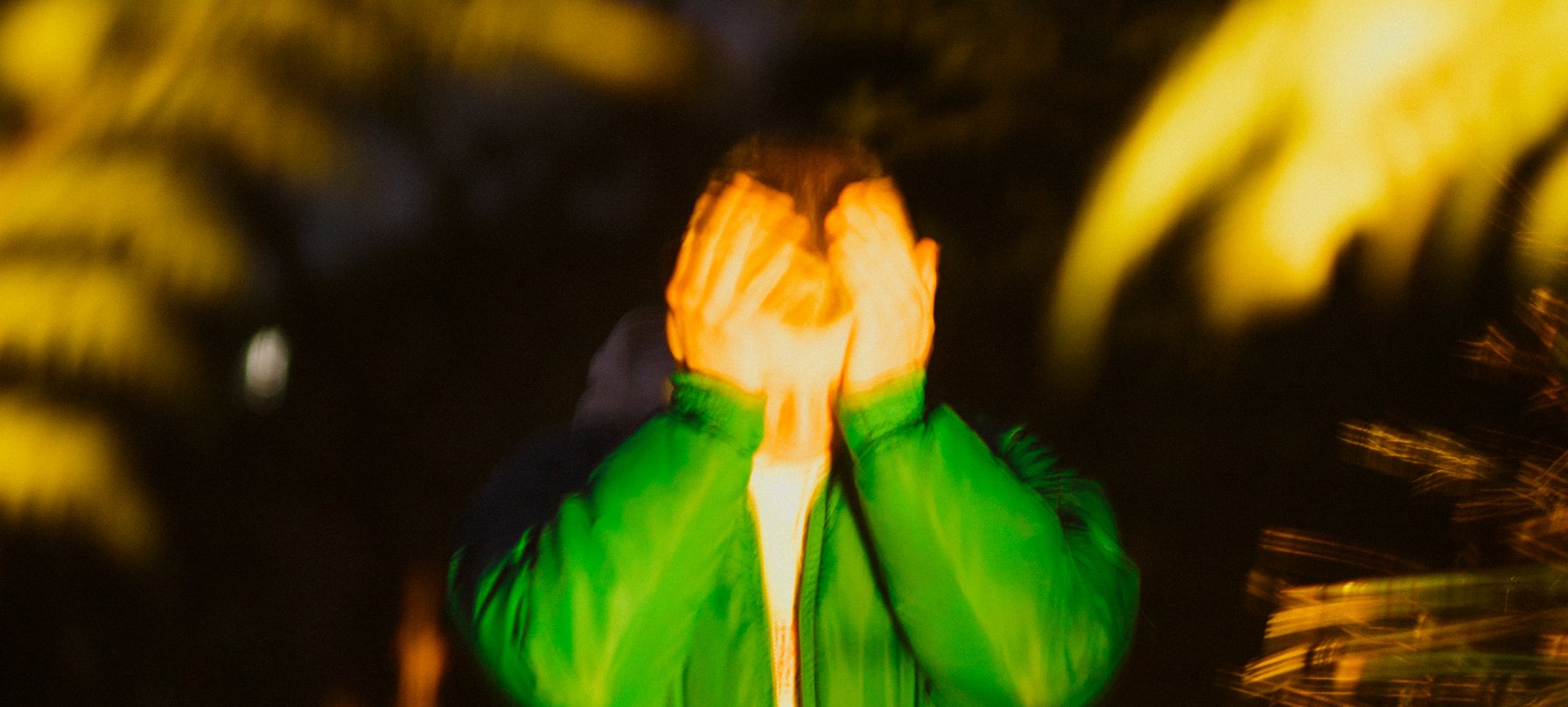 A photo of Godrich standinh amongst tall grass, wearing a bright green jacket and with their hands covering their eyes. Bright light warps this photo making the image look gliched. Credit Thomas Wood.