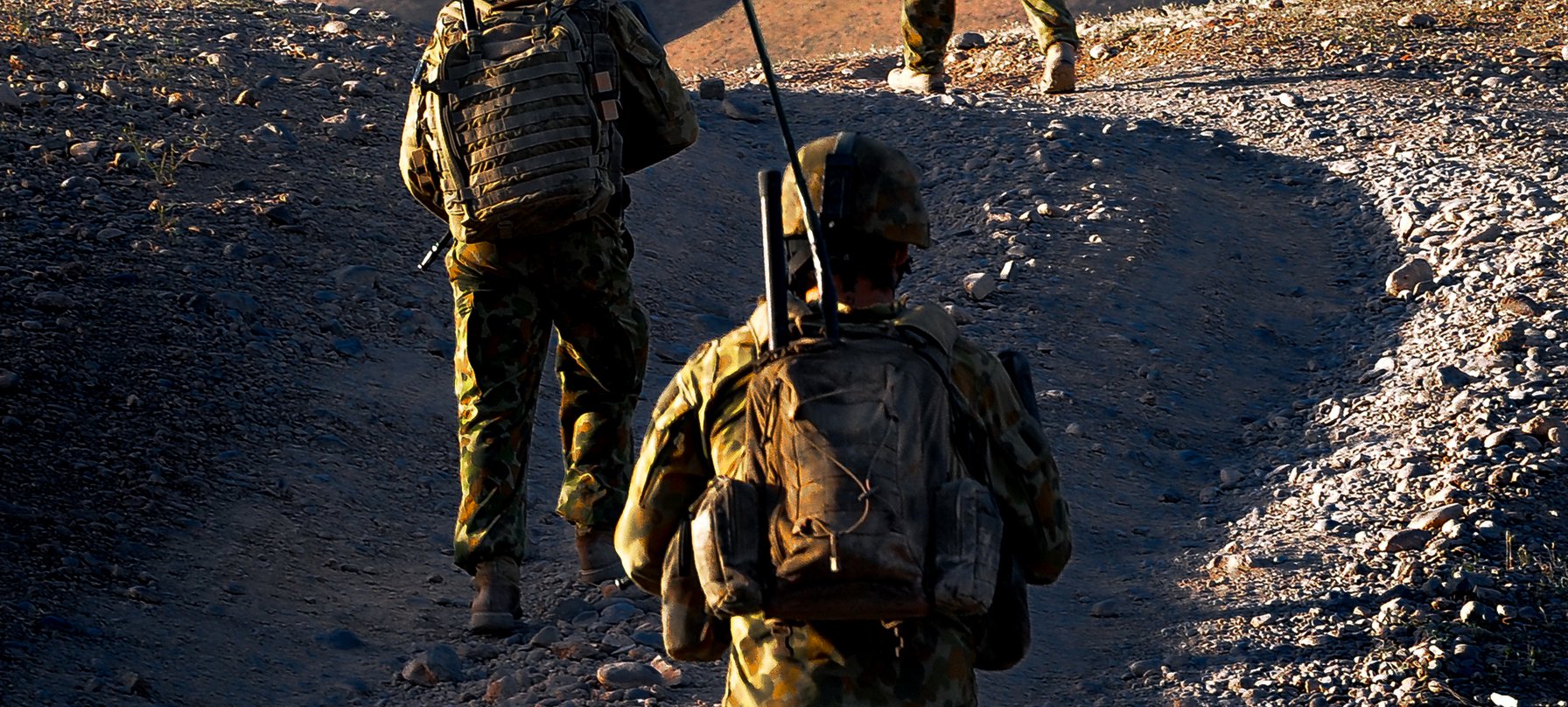 A photo of three people in modern army camouflage walking up a gravel hill. Each person wears a helmet and carries a backpack with a tall antenna pointing straight up. Image Mark Direen.