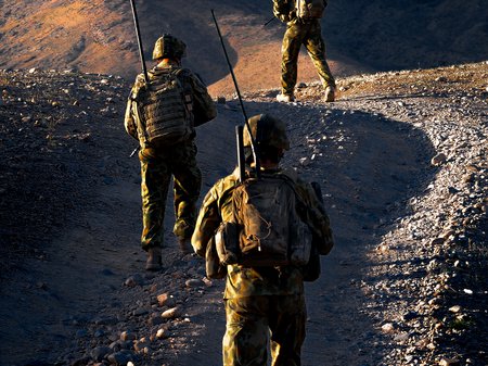A photo of three people in modern army camouflage walking up a gravel hill. Each person wears a helmet and carries a backpack with a tall antenna pointing straight up. Image Mark Direen.