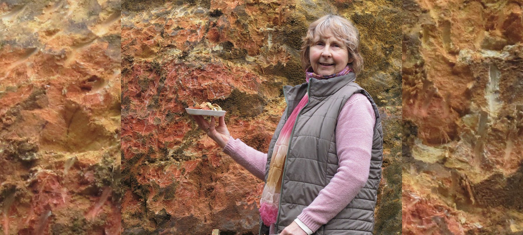 A photo of Chris T Wilson standing in front of a fiery-red and orange rock wall. Chris has a small pick axe in one hand and is holding a collection of rock fragment in the other. Chris smiles at the camera, looking proud of her haul. Credit Chris T Wilson