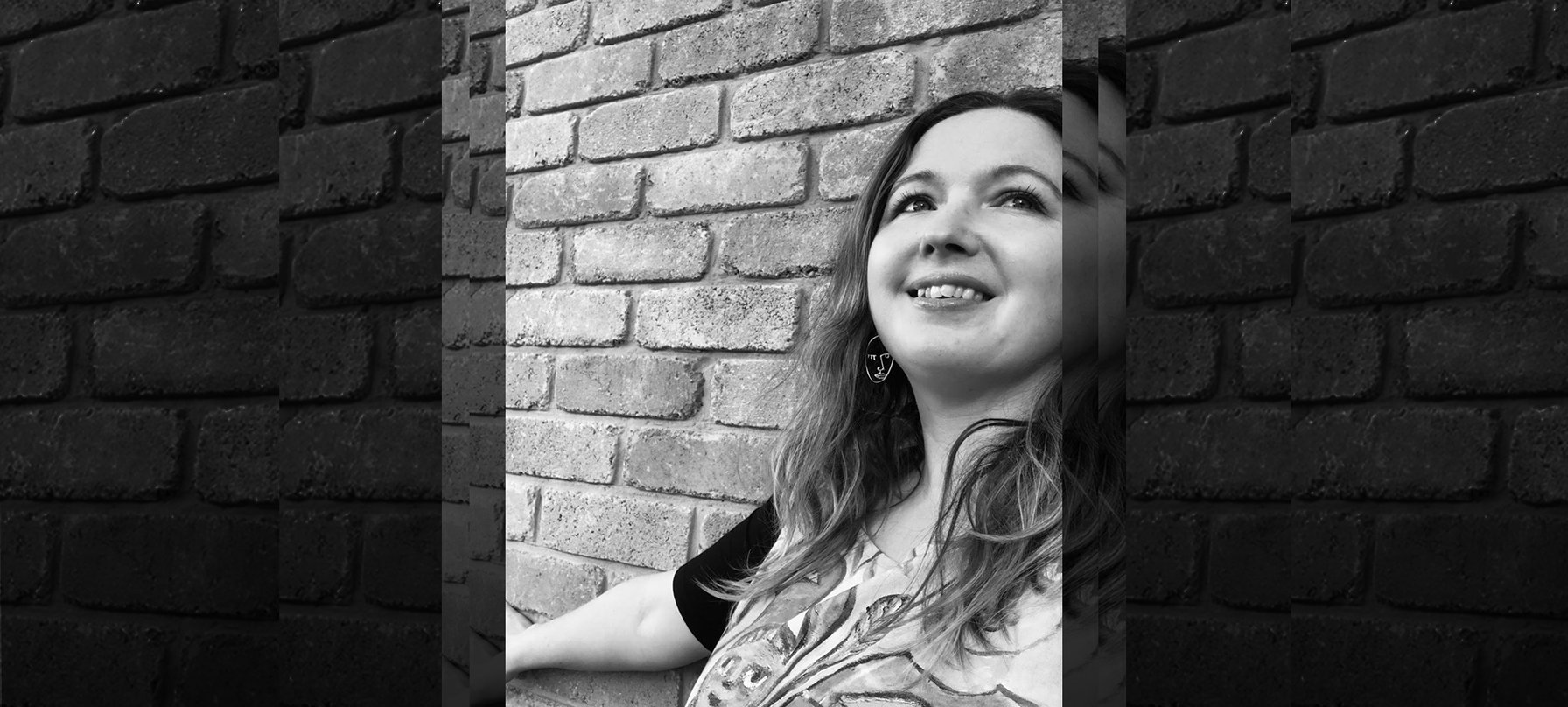 A black and white photo of Hayley Strutt smiling with her back against a brick wall. Hayley has pale skin and long brown hair. Credit Hayley Strutt.