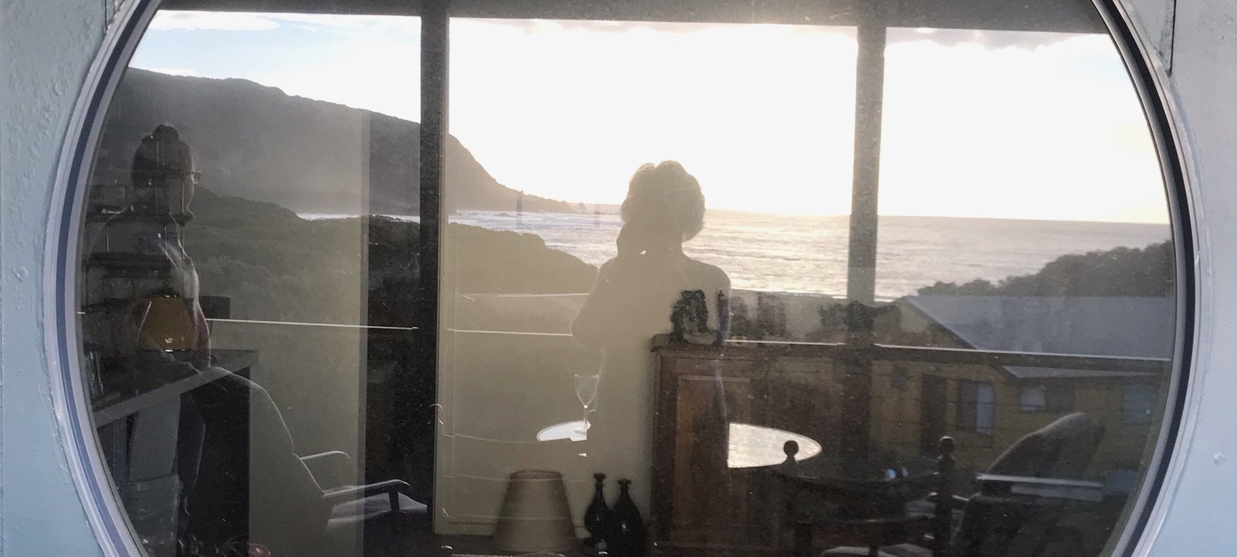 A photo taken in the reflection of a large round window. The reflection captures a sea-side scape of flat water, coastal hills and a sunlit sky. Credit Irene Briant.