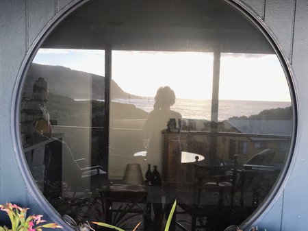 A photo taken in the reflection of a large round window. The reflection captures a sea-side scape of flat water, coastal hills and a sunlit sky. Credit Irene Briant.