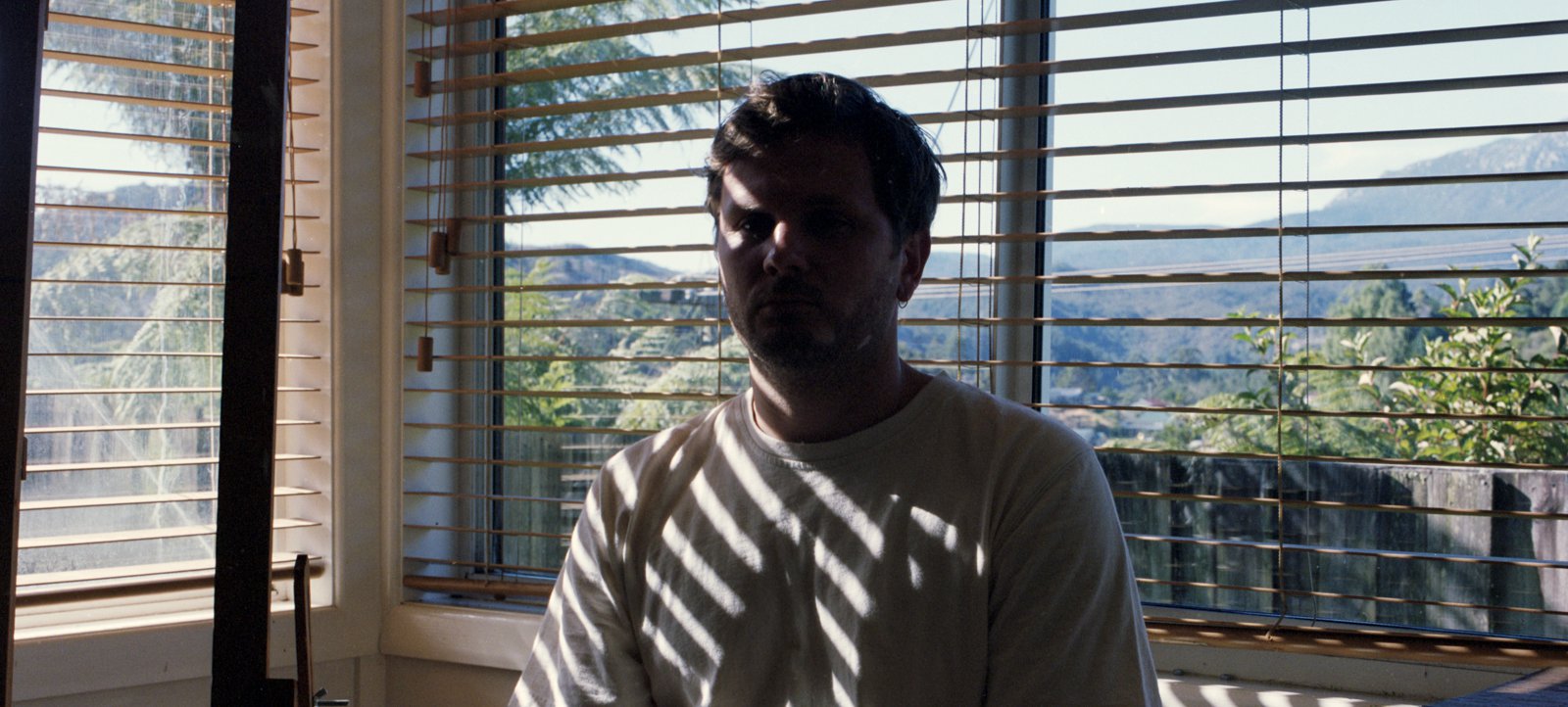 A photo of Carl Ross sitting on a wooden chair in a sunny room. Carl's face is blocked by shade, he wears a white t-shirt and tan pants. Venetian blinds cover all windows and mark Carl and the surrounding room with diagonal stripes. Credit Noah Thompson.