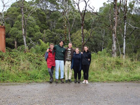 A photo of 5 UTAS Architecture students standing in a row infront of a dense and wet Queenstown bushscape. The have their arms around each others shoulders and are all smiling wide at the camera. Trees in the background are similarly tangled. No credit.