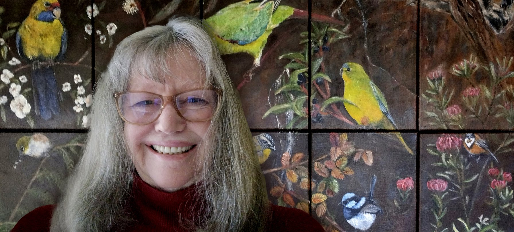 A photo of Maureen Bennetts smiling broadly in front of her artwork. Maureen's work is a series of smaller canvases that create a large picture when put together. The painting peeks into a scene of bright birds, waratah flowers and fagus leaves. No credit