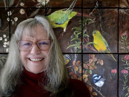 A photo of Maureen Bennetts smiling broadly in front of her artwork. Maureen's work is a series of smaller canvases that create a large picture when put together. The painting peeks into a scene of bright birds, waratah flowers and fagus leaves. No credit