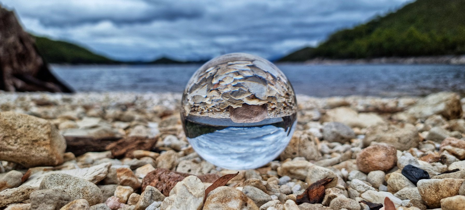 A photography artwork by Simon Geason. The shot is of a pale rock beach with a lake and cloudy blue sky in the background. Upon the rocks is a clear marble that is reflecting the scene within its body but completely upside down. Credit Simon Geason.