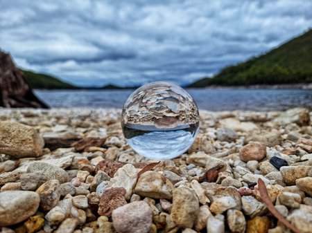 A photography artwork by Simon Geason. The shot is of a pale rock beach with a lake and cloudy blue sky in the background. Upon the rocks is a clear marble that is reflecting the scene within its body but completely upside down. Credit Simon Geason.