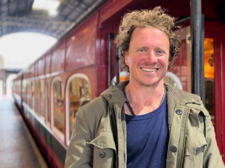 Artist Pete Hammond stands at a train station in front of a red train, smiling as he looks directly into the camera.