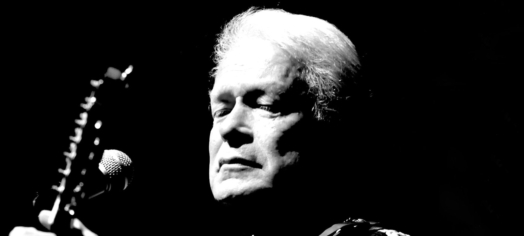 A black and white photo of Keith Potger performing with his guitar. Keith wears an intricutely embroidered Western shirt, has white hair and pale skin. He looks down at the strings of his guitar as he plays. Credit