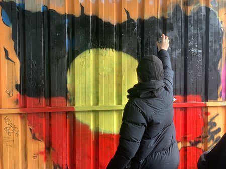 A photo of a person spray painting an artwork of an aboriginal flag.