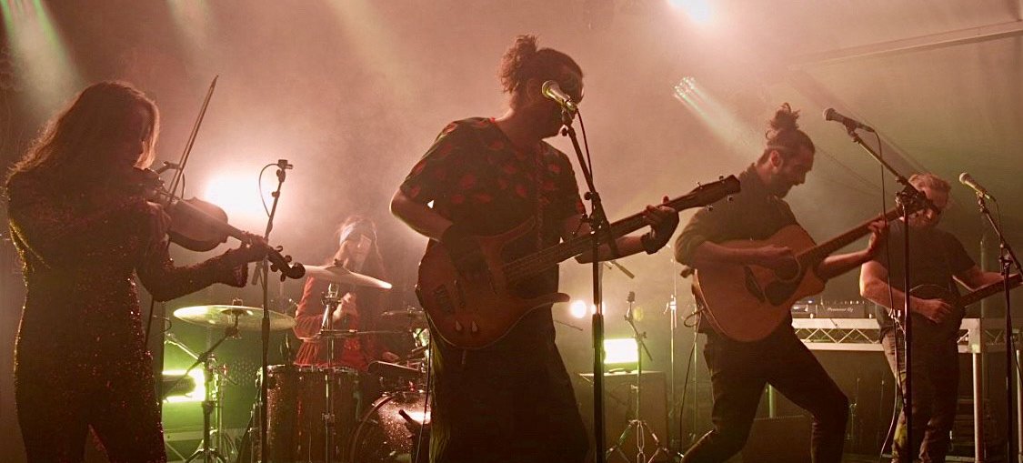 A photo of 5 band members of Alegría on a darkened stage with red and green lights shining from behind. The band play drums, an electric guitar, bass guitar, banjo and fiddle. Credit courtesy of artists.