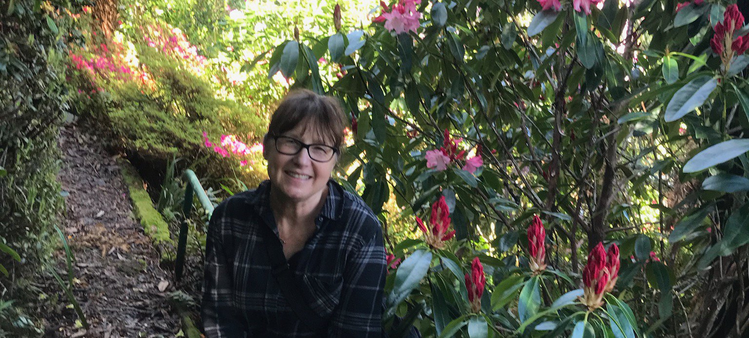 a photo of Cheryl Weare on a pathway covered in leaves and overflowing with green trees and vibrant pink flowers. Cheryl leans on a railing and smiles at the camera. Cheryl has pale skin and a brown full fringe with hair tied back. Credit artist.