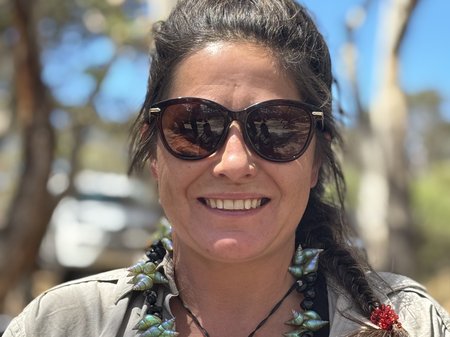 Krystelle Jordan stands in a sunny, tree-filled setting, wearing sunglasses and a shell necklace while smiling at the camera.