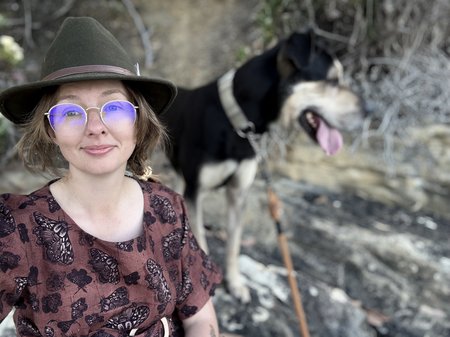 Artist Chelsea Smith sits outdoors on a rock, taking a selfie with branches and a dog softly blurred in the background.