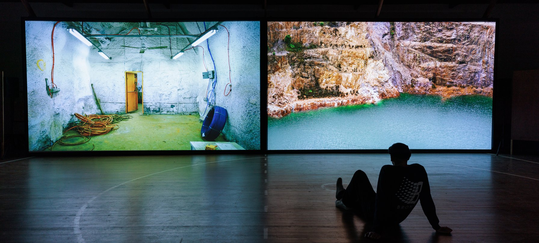 Person sits on the ground in the dark, watching a film split onto two screens.