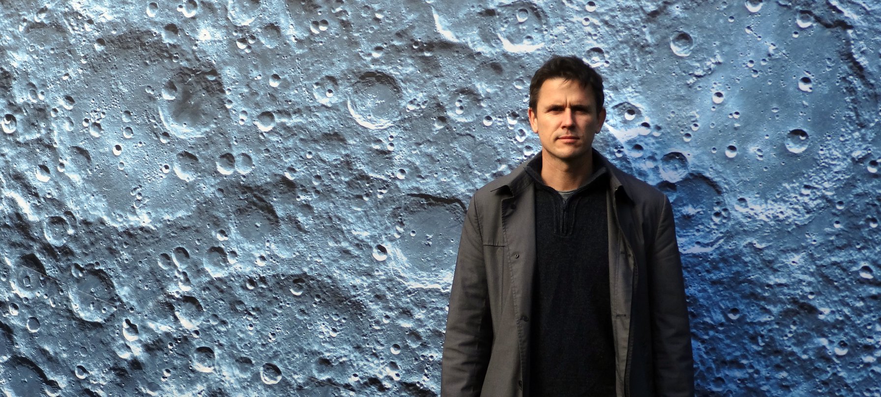 A photo of artist Luke Jerram standing in front of a large image of the surface of the moon. Photo courtesy of Luke Jerram.