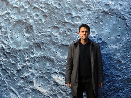 A photo of artist Luke Jerram standing in front of a large image of the surface of the moon. Photo courtesy of Luke Jerram.