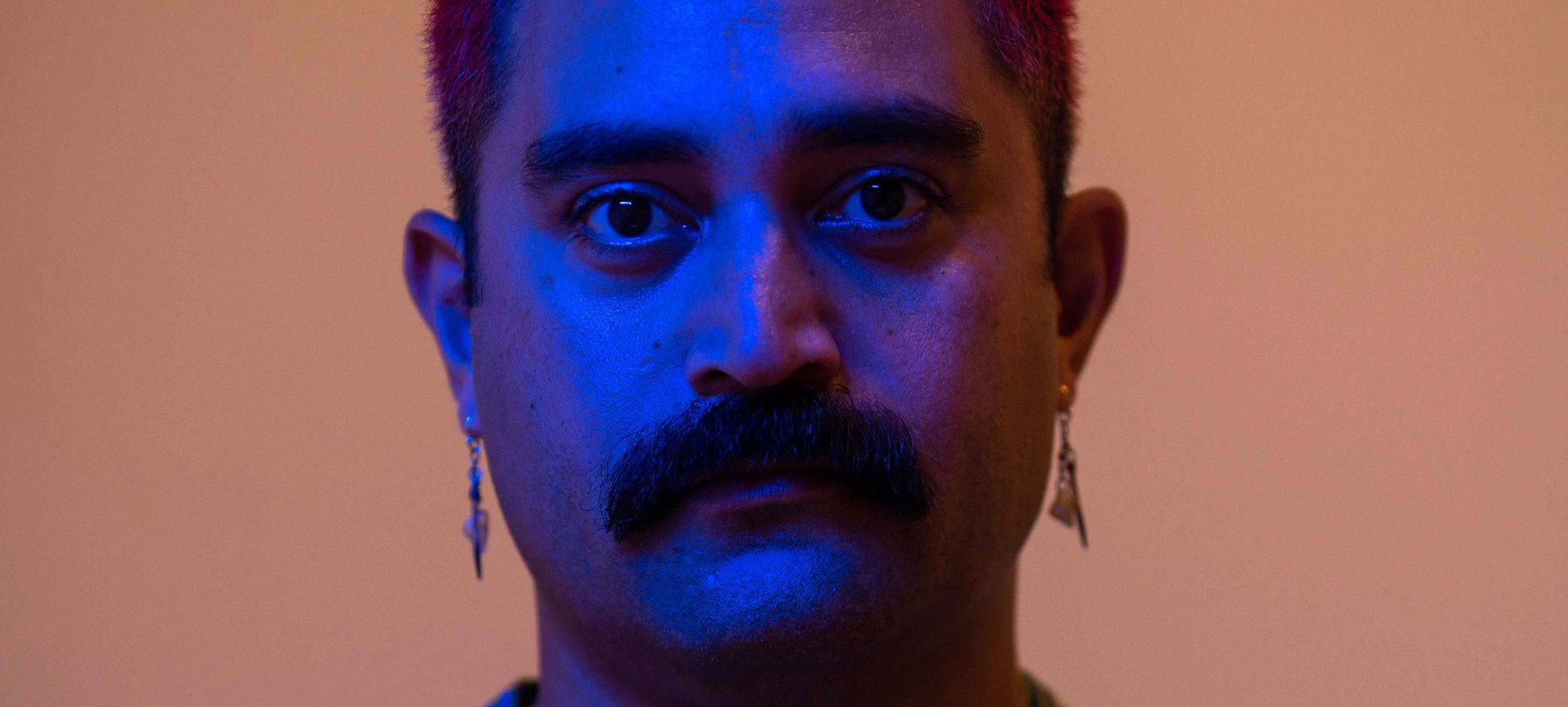 A photo of Léuli Eshrāghi looking at the camera with a dusty pink back ground and a blue light over their face. Léuli has tanned skin, red-dyed hair quiffed atop their head and a moustache that most would envy the lushness of. Credit Rhett Hammerton.