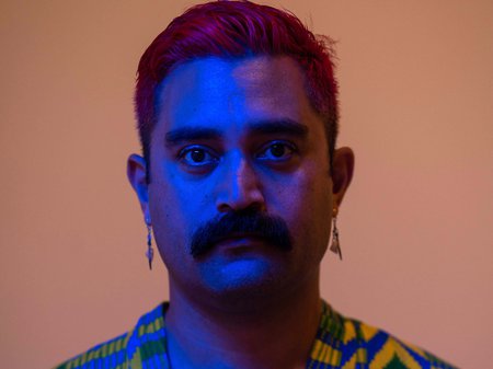 A photo of Léuli Eshrāghi looking at the camera with a dusty pink back ground and a blue light over their face. Léuli has tanned skin, red-dyed hair quiffed atop their head and a moustache that most would envy the lushness of. Credit Rhett Hammerton.