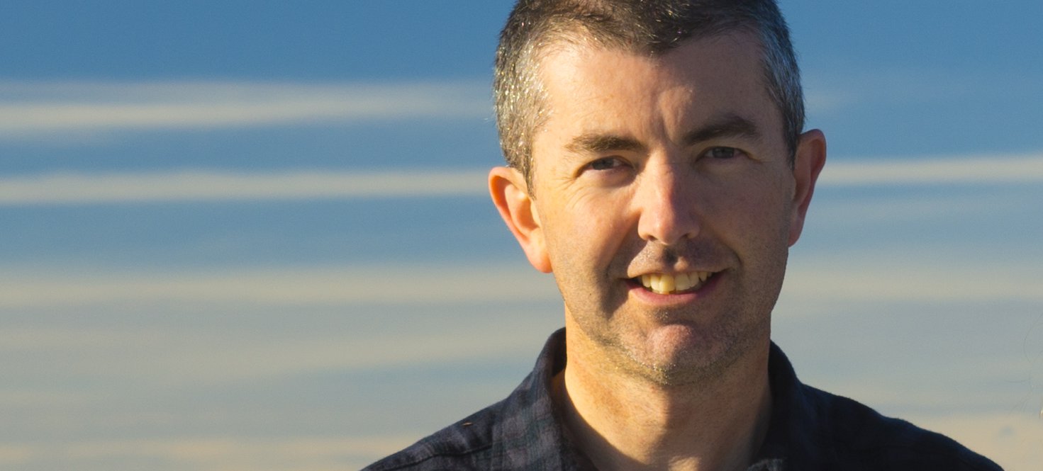 A photo of Mat James facing the camera and smiling. Mat wears a dark shirt and has pale skin, a clean-shaven face and a dark buzz-cut. Behind Mat is a blue sky streaked with long horizontal white clouds. Credit Mat James.