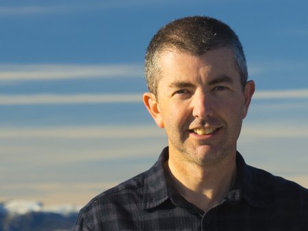 A photo of Mat James facing the camera and smiling. Mat wears a dark shirt and has pale skin, a clean-shaven face and a dark buzz-cut. Behind Mat is a blue sky streaked with long horizontal white clouds. Credit Mat James.