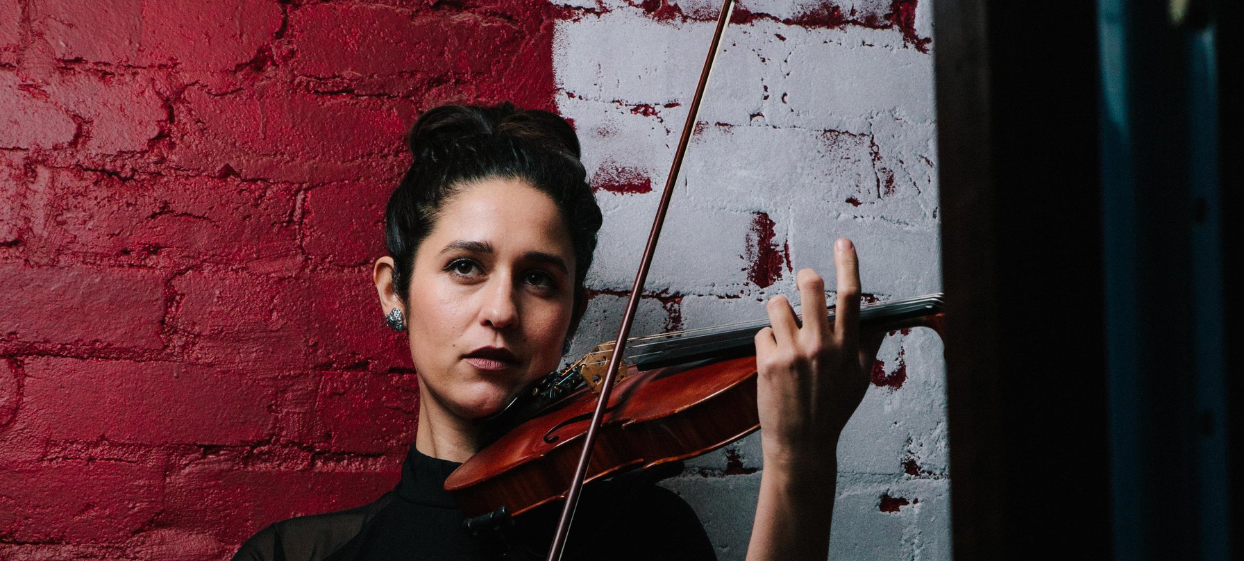 A photo of Natalya Bing playing her violin in front of a red and white painted brick wall. Credit Tom Wilkinson.