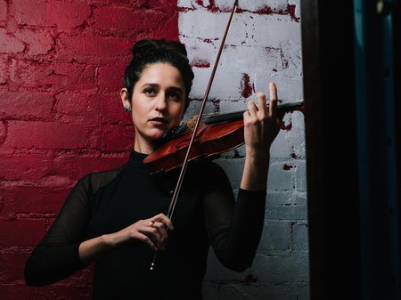 A photo of Natalya Bing playing her violin in front of a red and white painted brick wall. Credit Tom Wilkinson.