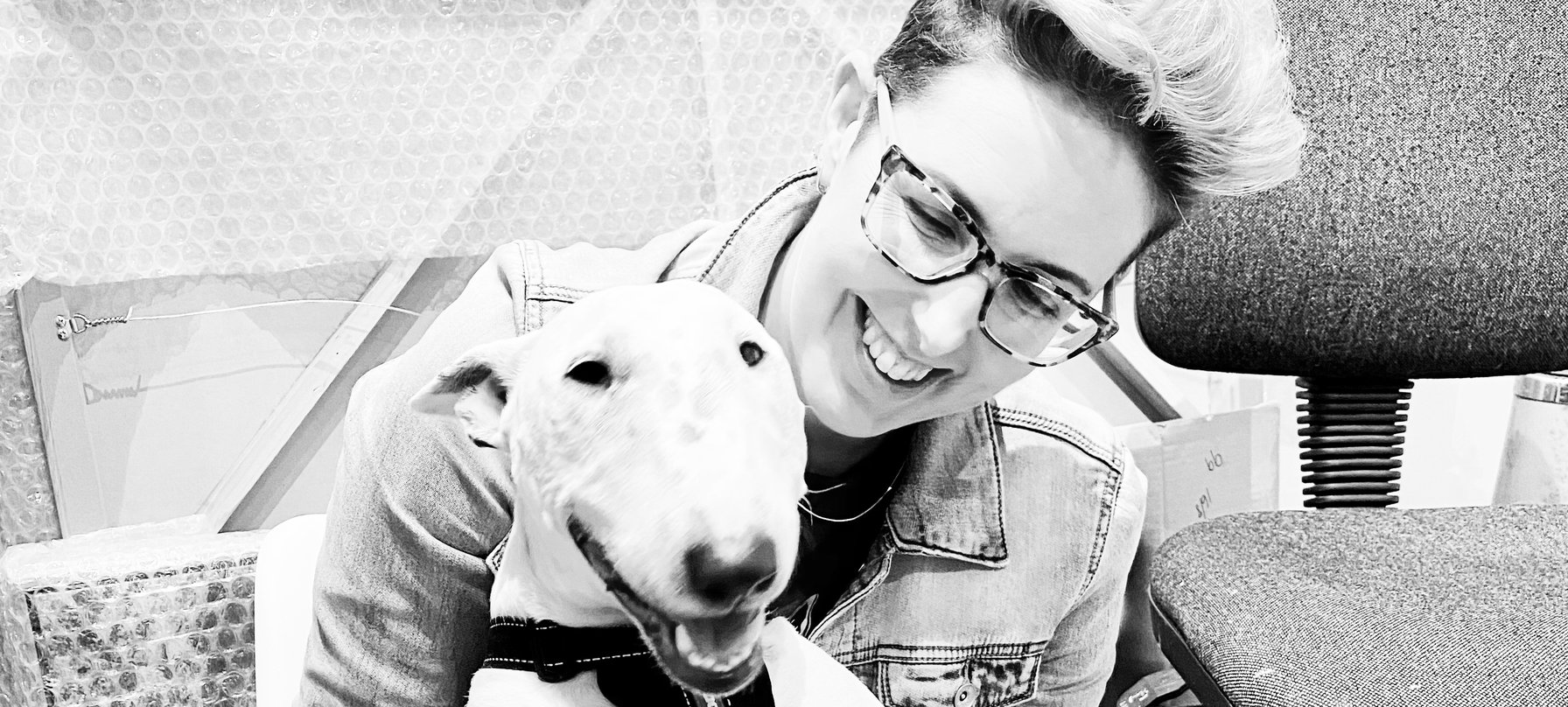 A black and white photo of Nicole Hutchins holding a bull terrier dog. Both appear to be smiling. Nicole has pale skin, wears wide-framed tortoise shell glasses and has short hair in a quiff atop their head. Credit Nicole Hutchins.