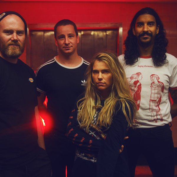 A photo of the 4 band members of Press Club. 4 people stand bunched together in a red room and with stern expressions on their faces. The closest person to the camera has their arms crossed against their chest. Credit Nick Manuell.