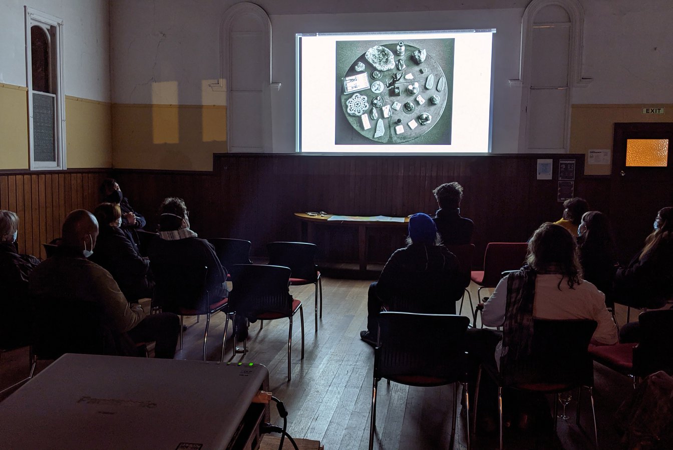 A photo of a dark hall interior, with a seated audience looking at an image projected onto the far wall. Photo courtesy of Lisa Sammut.